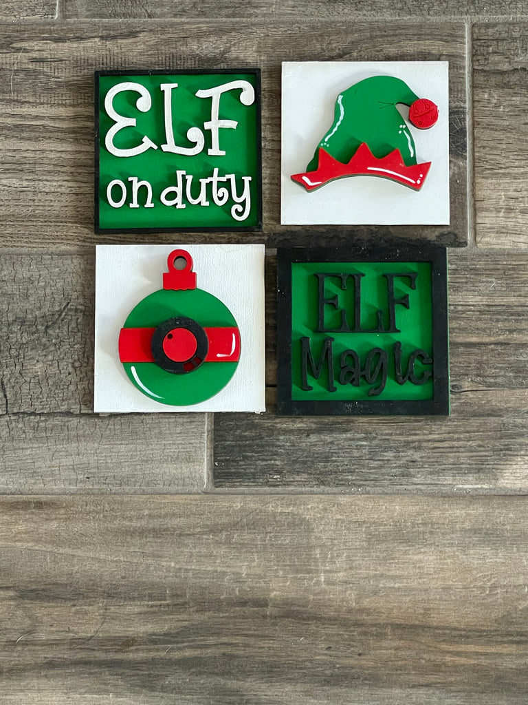 Four festive holiday-themed decorative wood items with elf motifs and phrases "elf on duty" and "elf magic," displayed leaning against a ladder on a wooden floor from Little August Ranch's Elf on Duty DIY - Leaning Ladder Insert Kit - Interchangeable Holiday Decor.