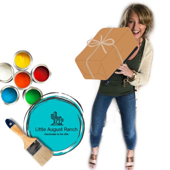 Woman holding a large cardboard cutout of the Summer Highland Cow Tiered Tray DIY Kit - Quick and Easy Tiered Tray Bundle, with paint cans, wood pieces, and a branded logo for Little August Ranch displayed around her.