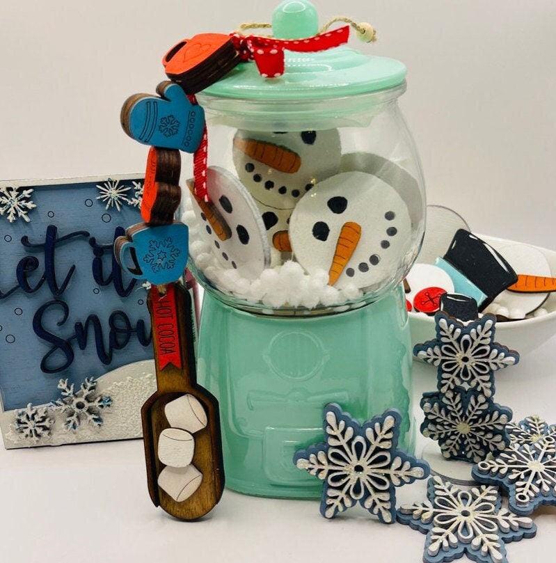 A green jar filled with Winter Gumball Machine Filler - DIY Gumball Filler Craft Kit by Little August Ranch, accompanied by wooden snowflake ornaments, a blue &quot;Let it snow&quot; sign, and a wooden spoon with marshmallow accents—all perfect elements for cr