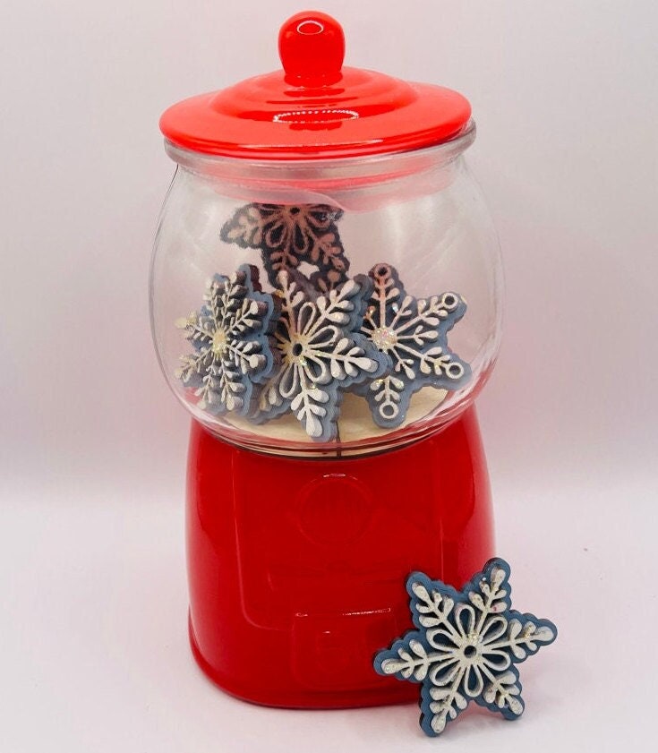 A red-lidded glass jar on a red base holds snowflake-shaped cookies with white icing, perfect for a holiday DIY kit. One cookie rests at the base of the jar. The Winter Gumball Machine Filler - DIY Gumball Filler Craft Kit by Little August Ranch is a