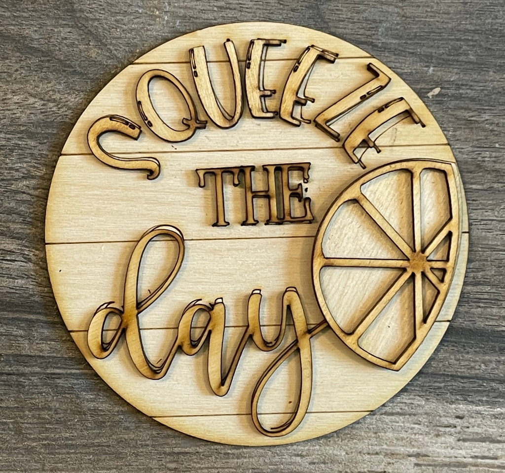 Wooden coaster engraved with the phrase &quot;Squeeze the day&quot; and an image of a lemon half, set on a gray wood surface from the Little August Ranch Lemon Tiered Tray DIY Paint Kit - Farmers Market Wood Blanks - U-Pick- Summer Paint Kit.