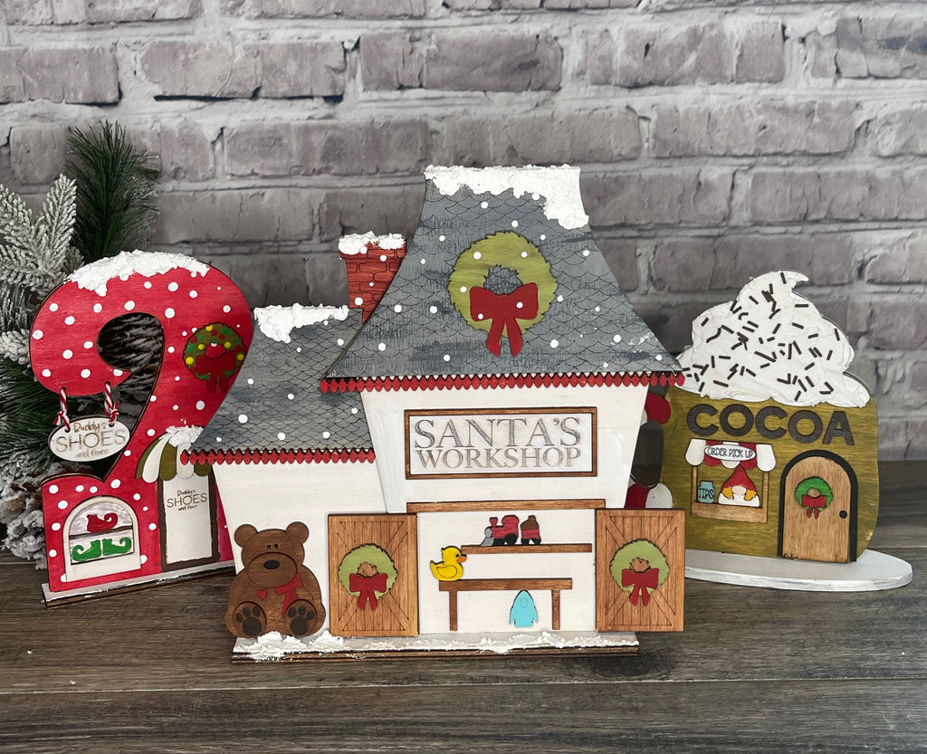 Holiday-themed decorative setup featuring a "Santa's workshop" house, a candy cane, and cocoa stand ornaments against a brick backdrop as part of a Little August Ranch Christmas Village display featuring Christmas Village Self Standing Double Sided Pieces - Cocoa Hut - Winter Village Wood Blanks.