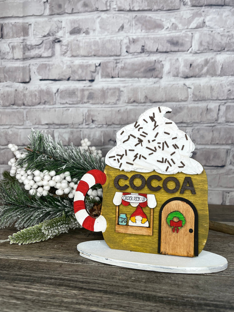 A decorative wooden Cocoa Hut stand from Little August Ranch, with a whipped cream design on top, displayed alongside a pine branch and berries on a wooden surface against a brick wall background, forms part of a charming DIY Village setup.