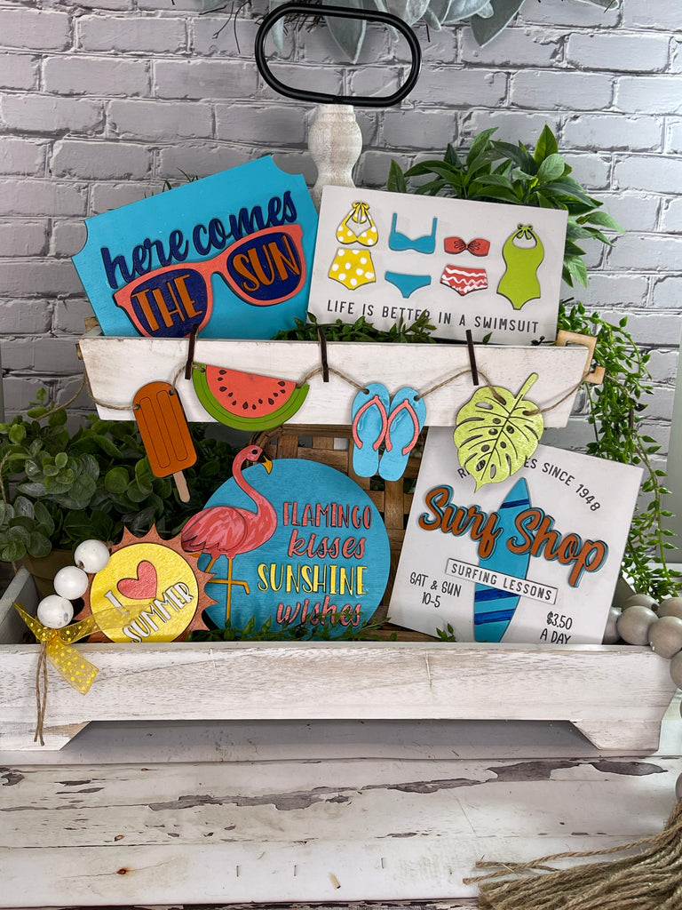 Assortment of colorful DIY Summer Beach Tiered Tray - Surf Shop Tier Tray Bundle signs and decorations from Little August Ranch displayed on a tiered tray with a brick backdrop.