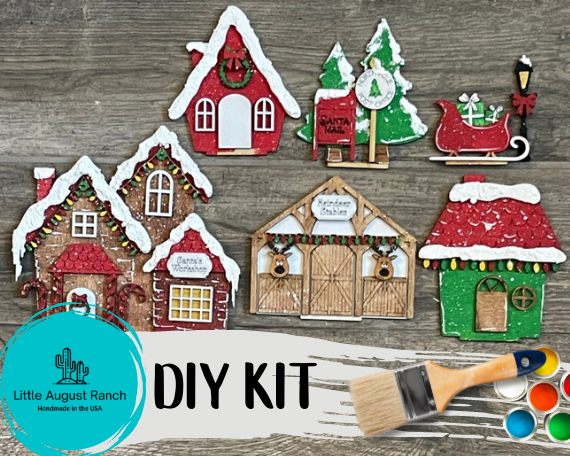 A Little August Ranch DIY Christmas Santas Village Standing Pieces - Winter Village - Christmas Shelf Decor Blank Kit - Reindeer Barn, Sleigh, Santa Mailbox Blank with wooden items and a paint brush.