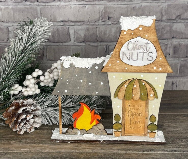 A DIY kit of Little August Ranch's Christmas Village Self Standing Double sided Pieces - Chestnut Roaster with a fireplace and pine cones.