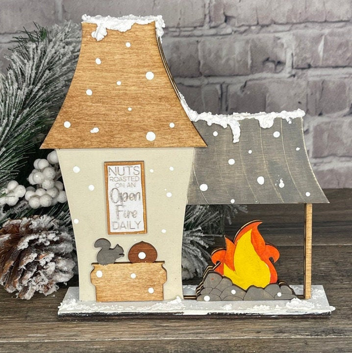 A wooden house with a fireplace, pine cones, and a Little August Ranch Christmas Village Self Standing Double sided Pieces - Chestnut Roaster.